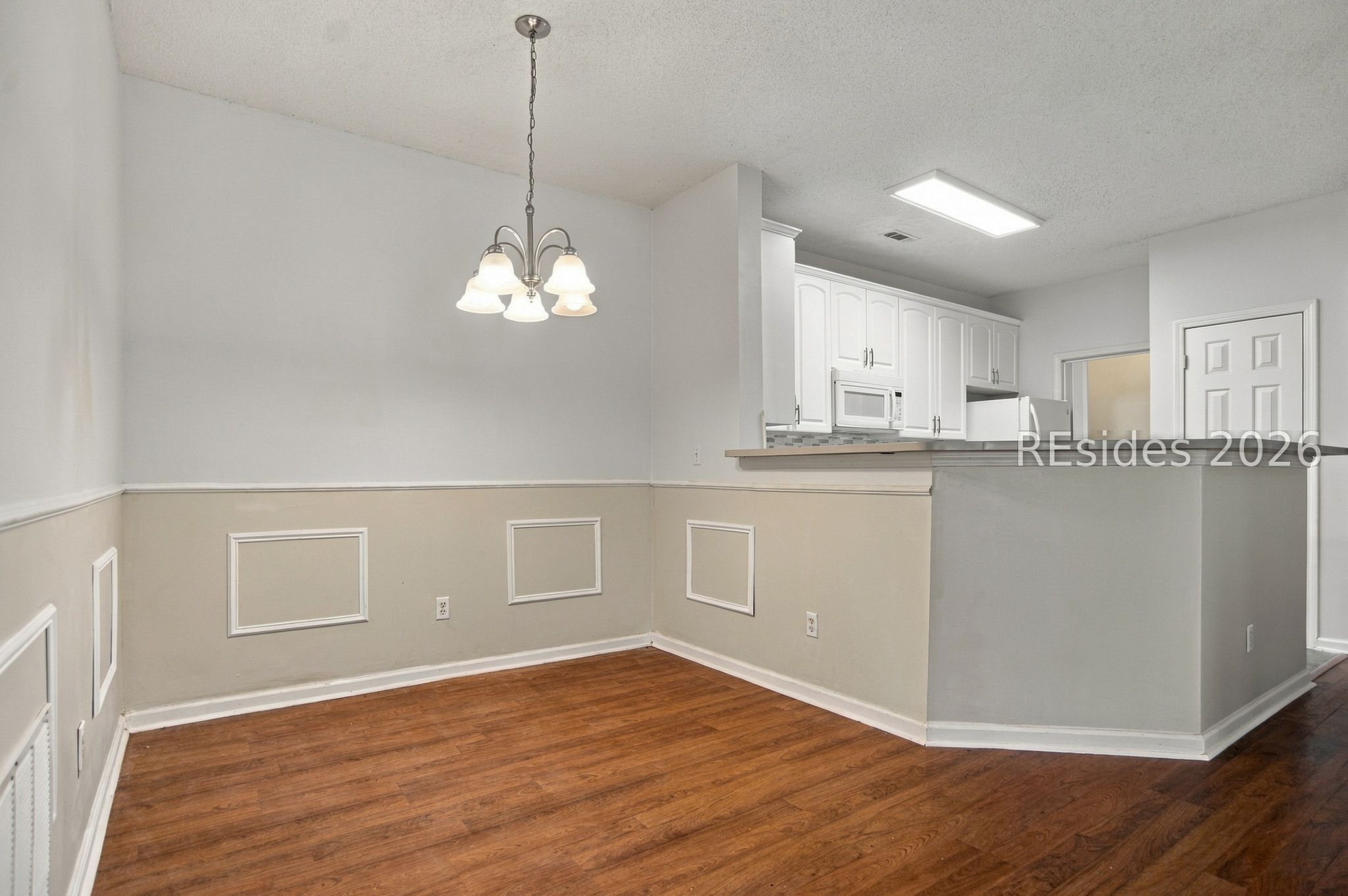 Empty dining area with hardwood floors, decorative wall trim, and an open kitchen with white cabinets.