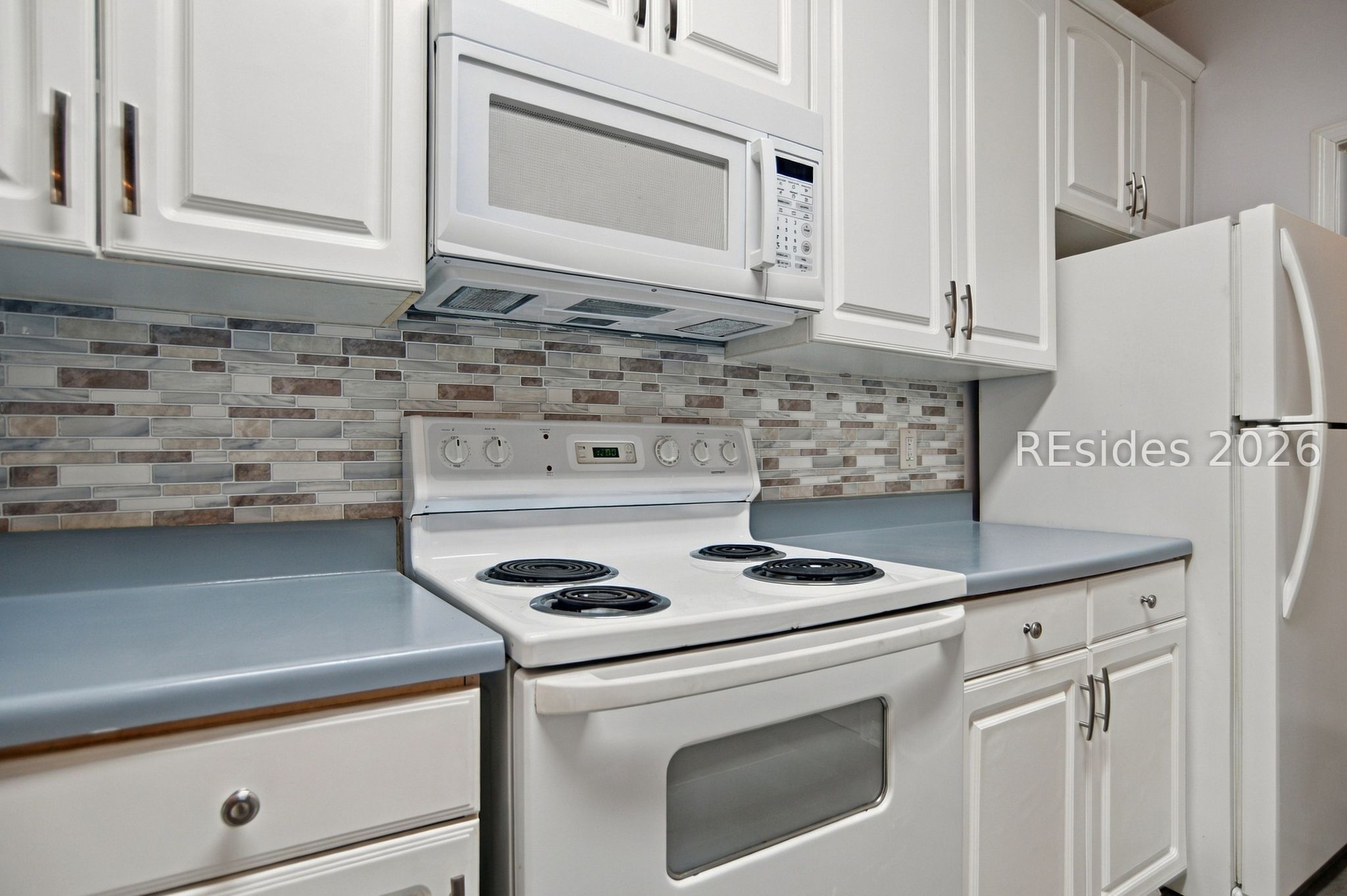 Kitchen with white cabinets, blue-gray counters, tile backsplash, electric range, microwave, and refrigerator.