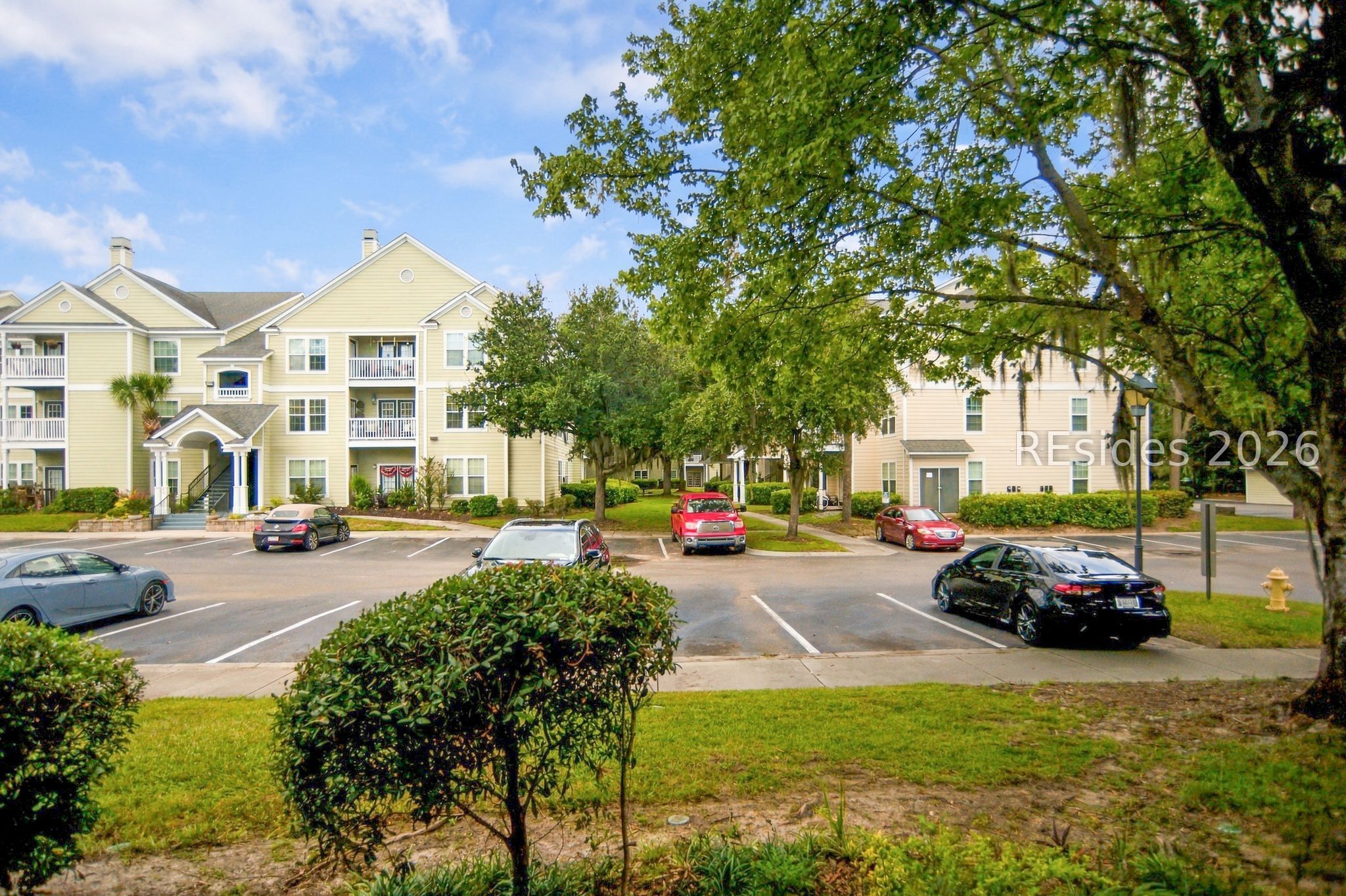 Pale yellow apartment building with balconies behind a tree-lined parking lot.