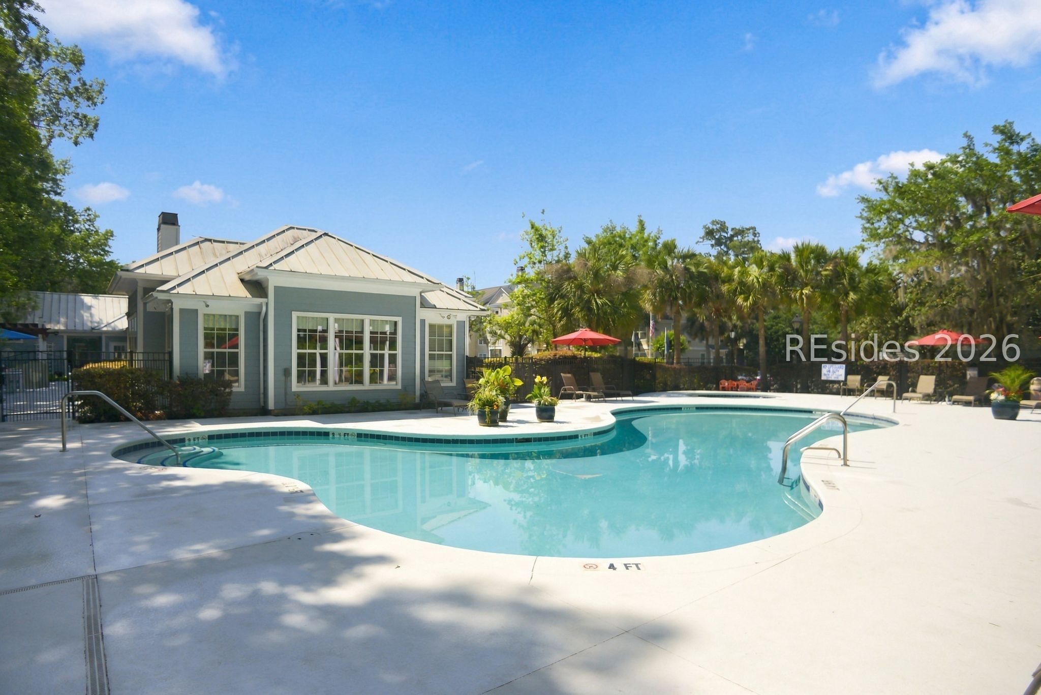 Community swimming pool beside a gray clubhouse, with palm trees and a red patio umbrella.