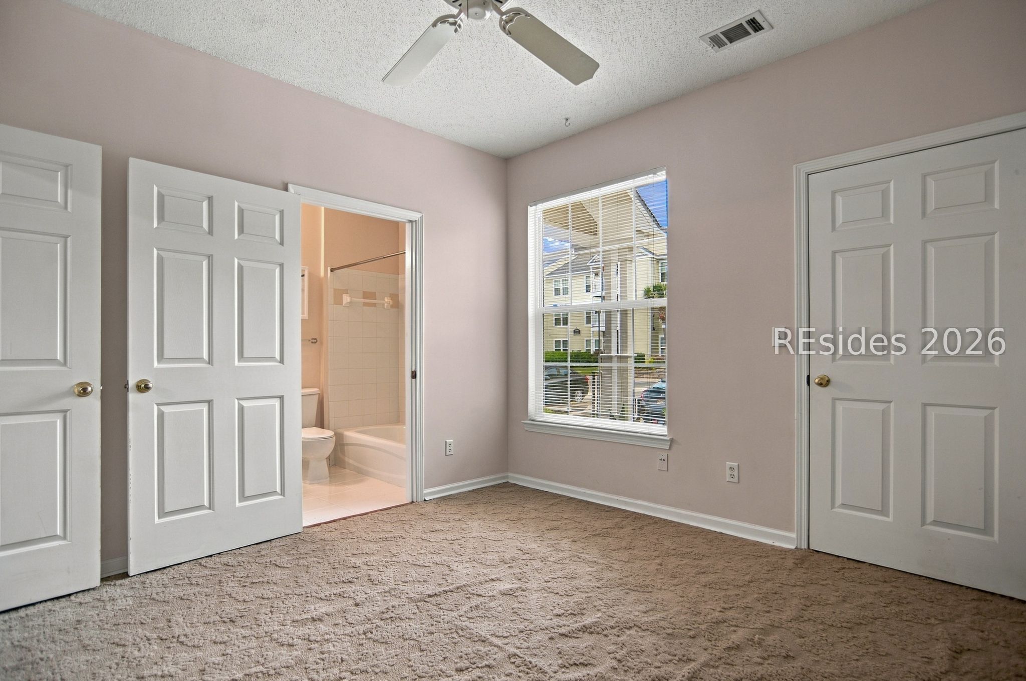 Empty bedroom with beige carpet, ceiling fan, and window overlooking a neighboring apartment building.