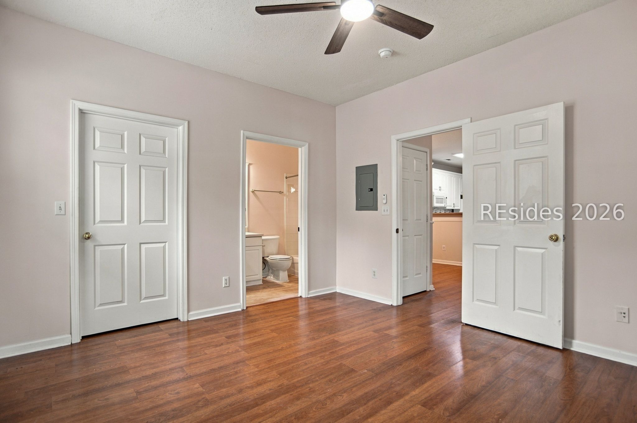Empty room with hardwood floors, ceiling fan, and open doorways to a bathroom and kitchen.