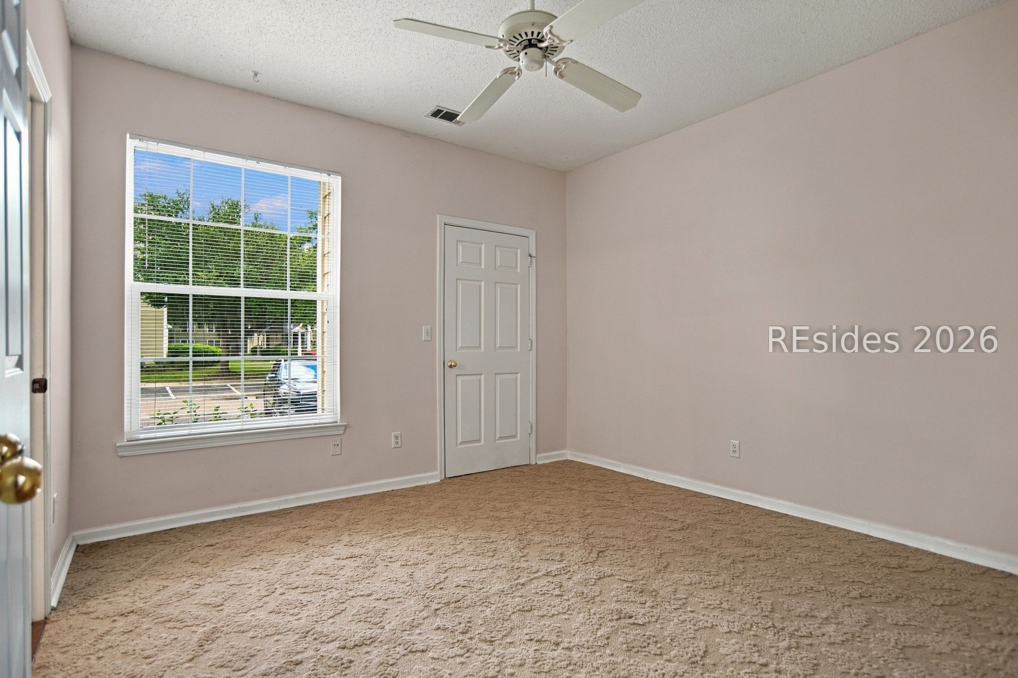 Empty carpeted bedroom with a ceiling fan and a large window overlooking greenery.
