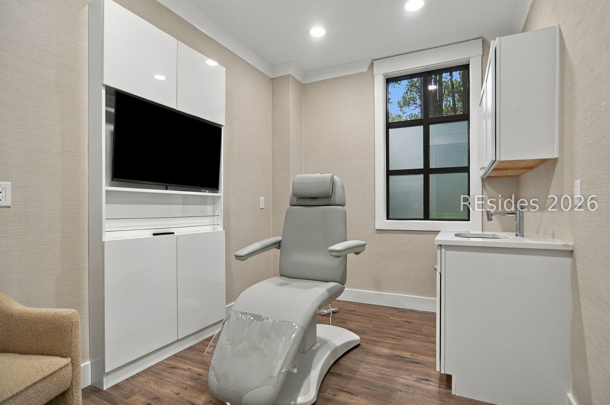 Treatment room with a reclining examination chair, built-in cabinetry, wall-mounted screen, and sink.
