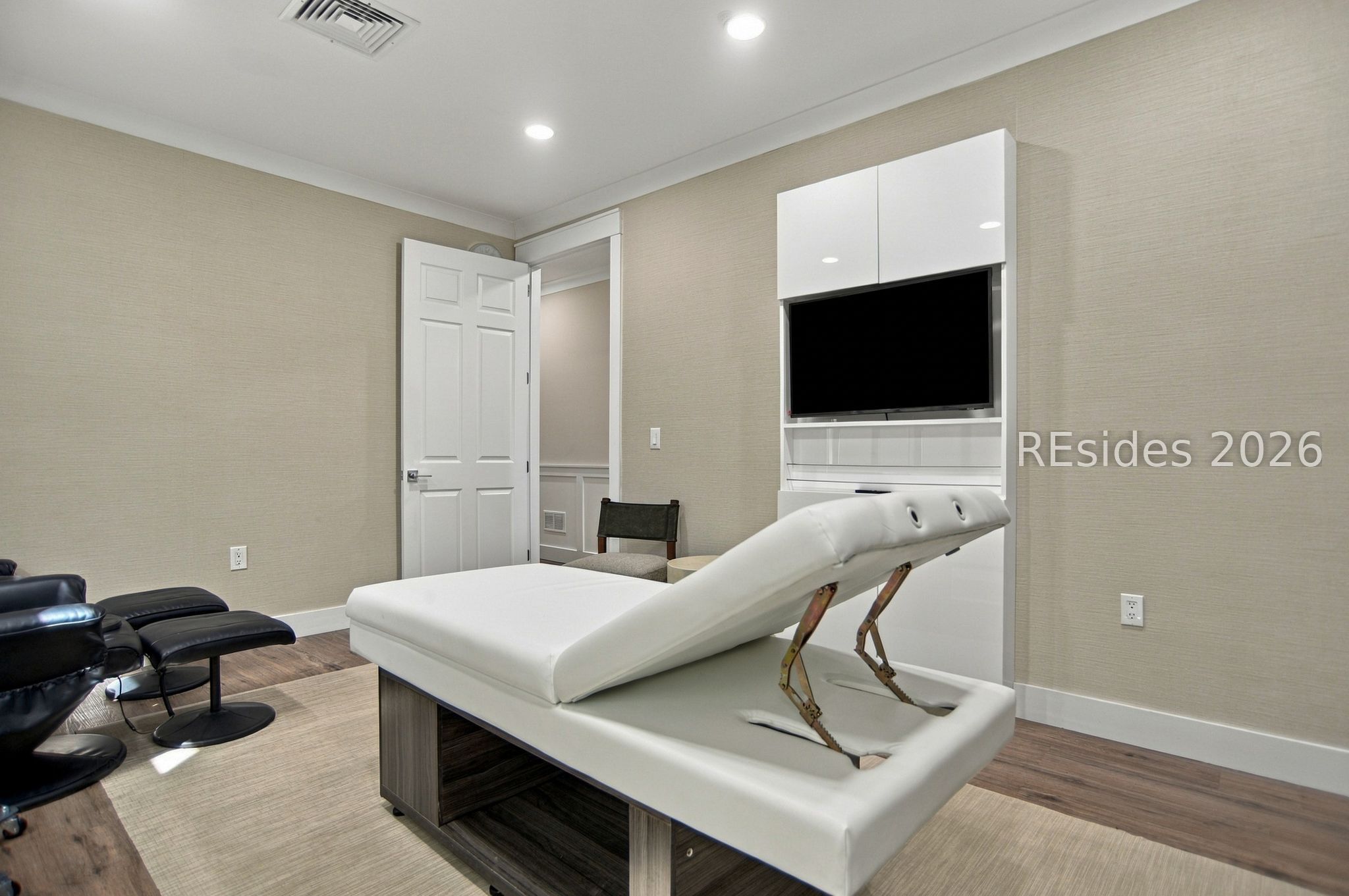 Furnished treatment room with an adjustable table, reclining chairs, and a wall-mounted television.