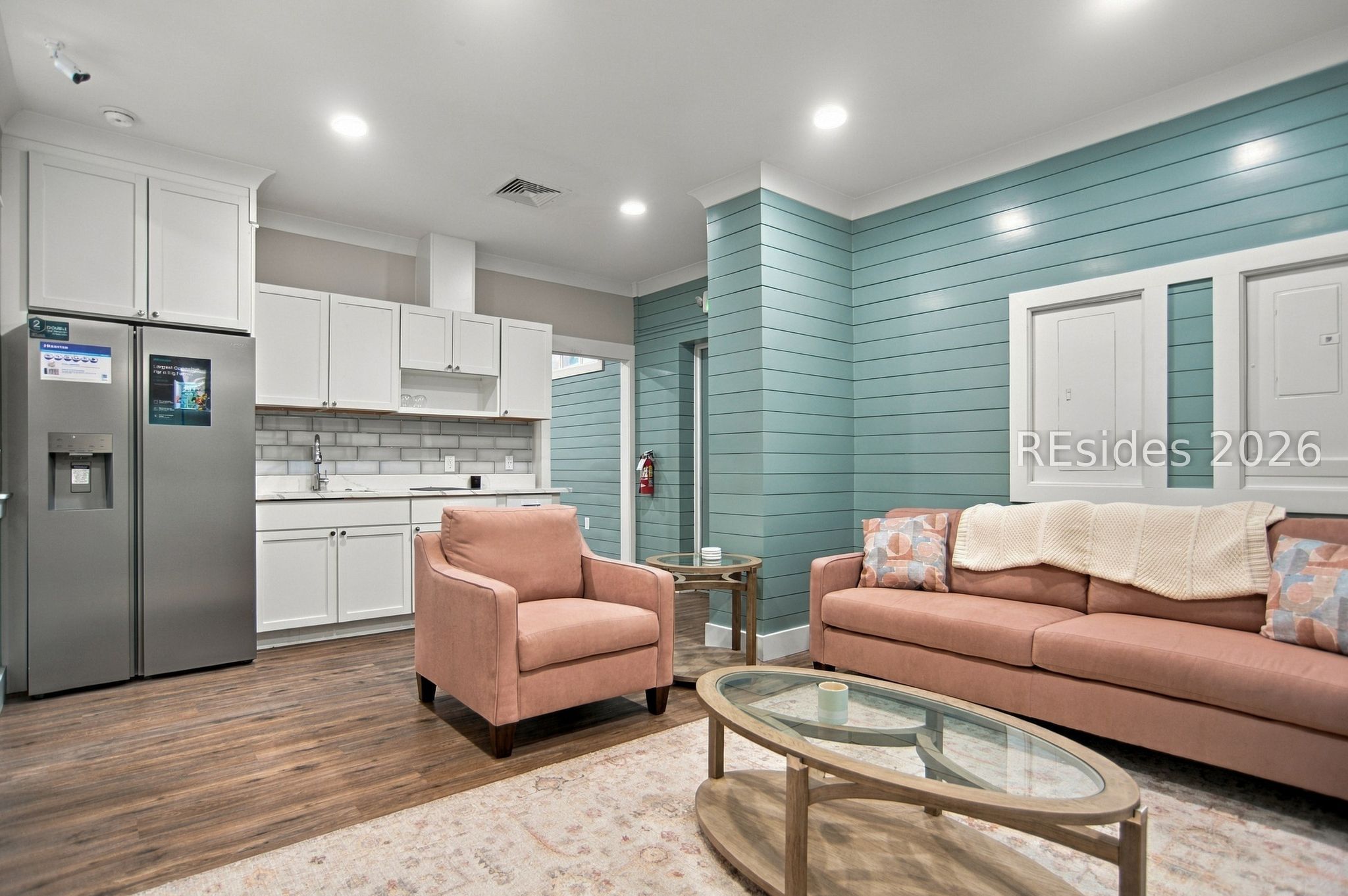 Open-plan living room and kitchen with teal shiplap walls, white cabinets, and pink sofa and armchair.
