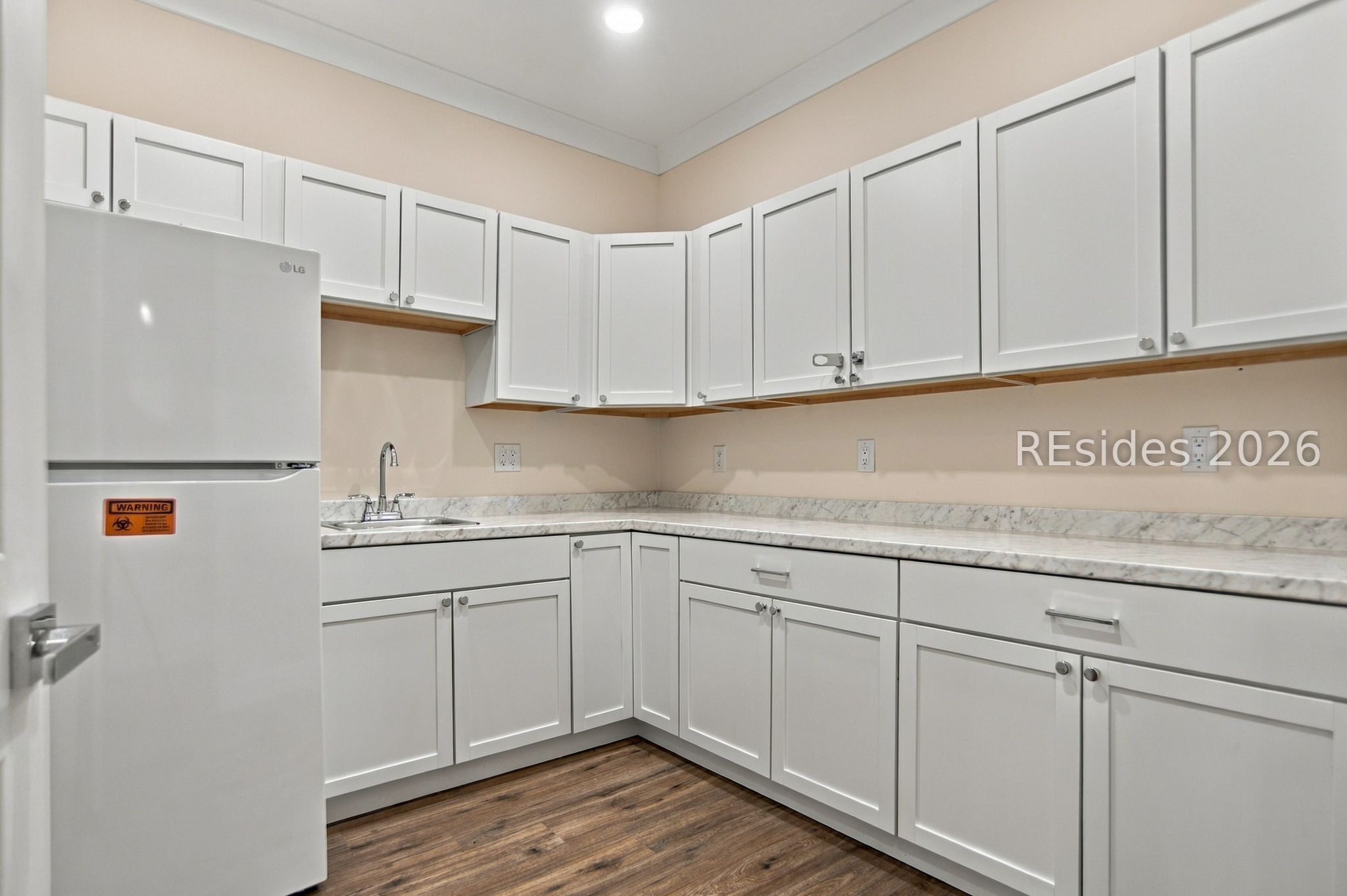 Vacant kitchen with white cabinetry, marble-pattern counters, a corner sink, and a white refrigerator.
