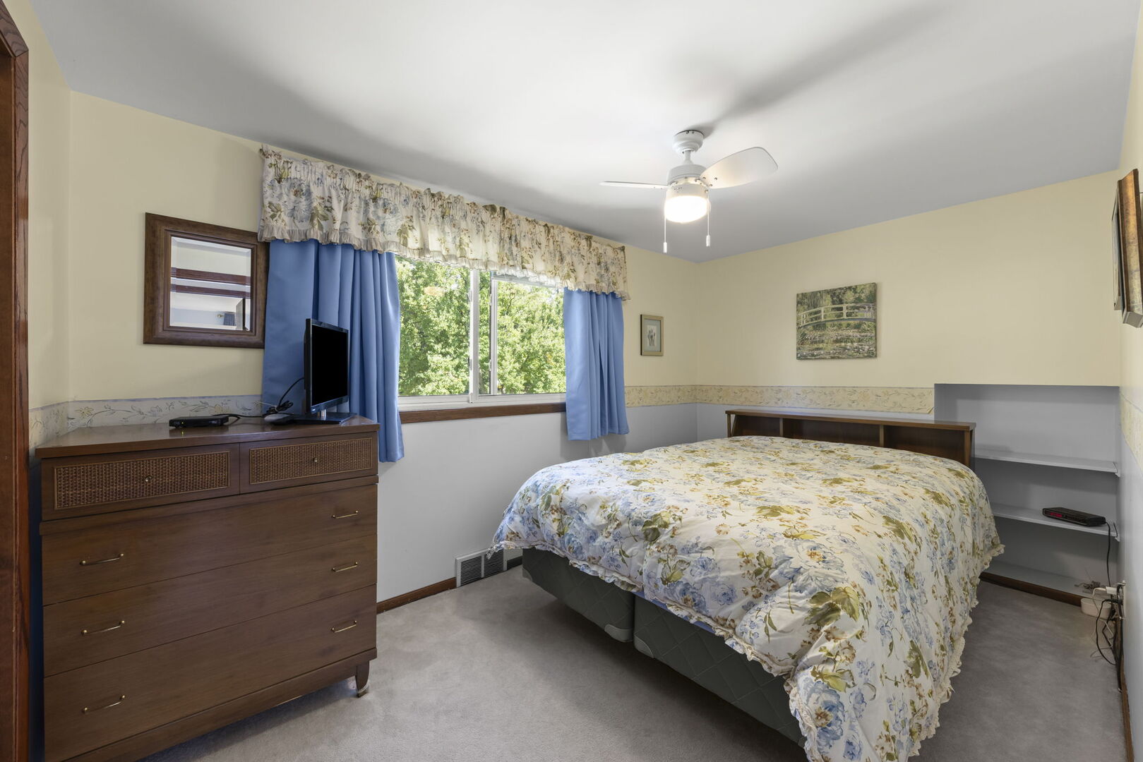 Bedroom with a floral bedspread, wooden dresser, and blue curtains framing a leafy view.