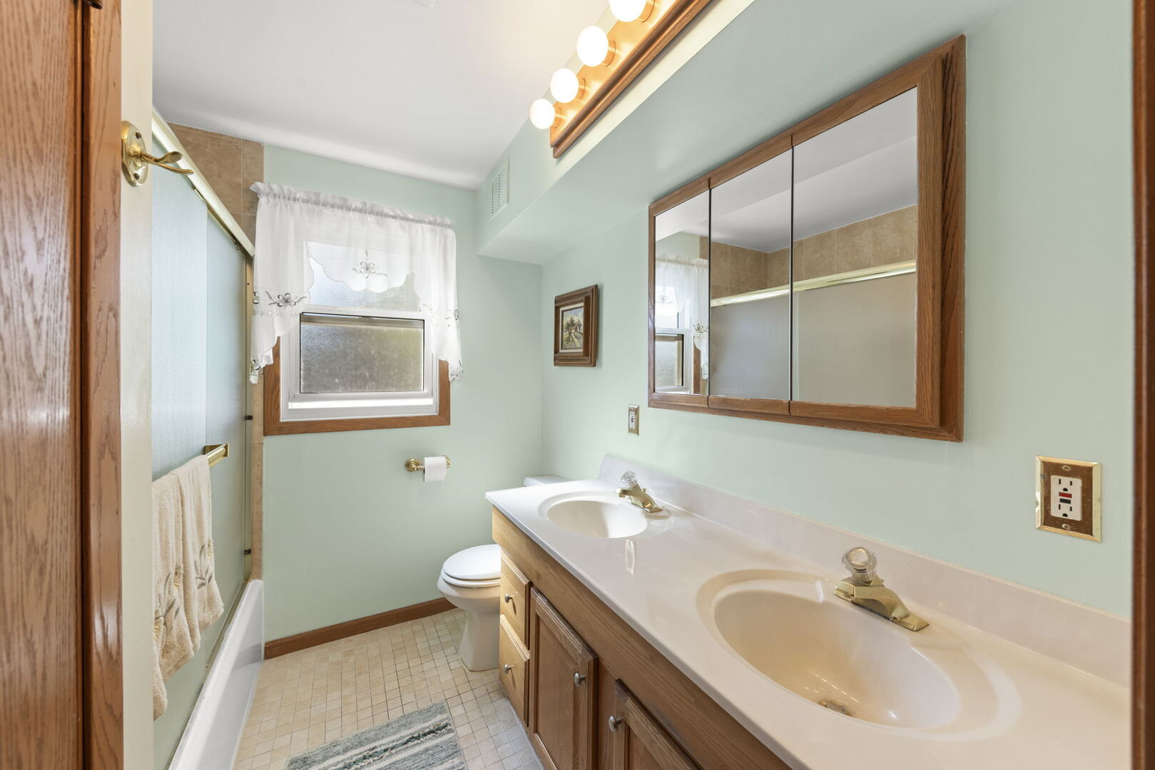 Bathroom with a double-sink wood vanity, mirrored cabinet, and tub-shower beside a window.