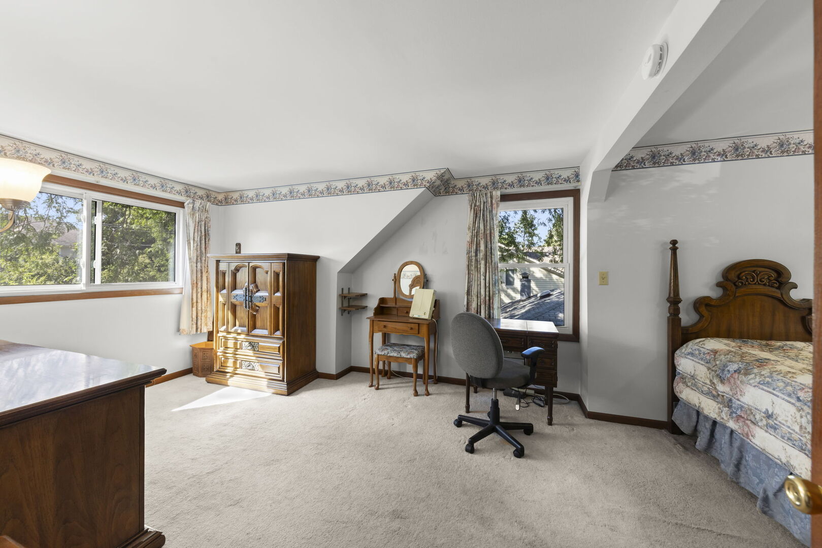 Furnished bedroom with a wooden armoire, vanity, desk, and bed beneath a sloped ceiling.