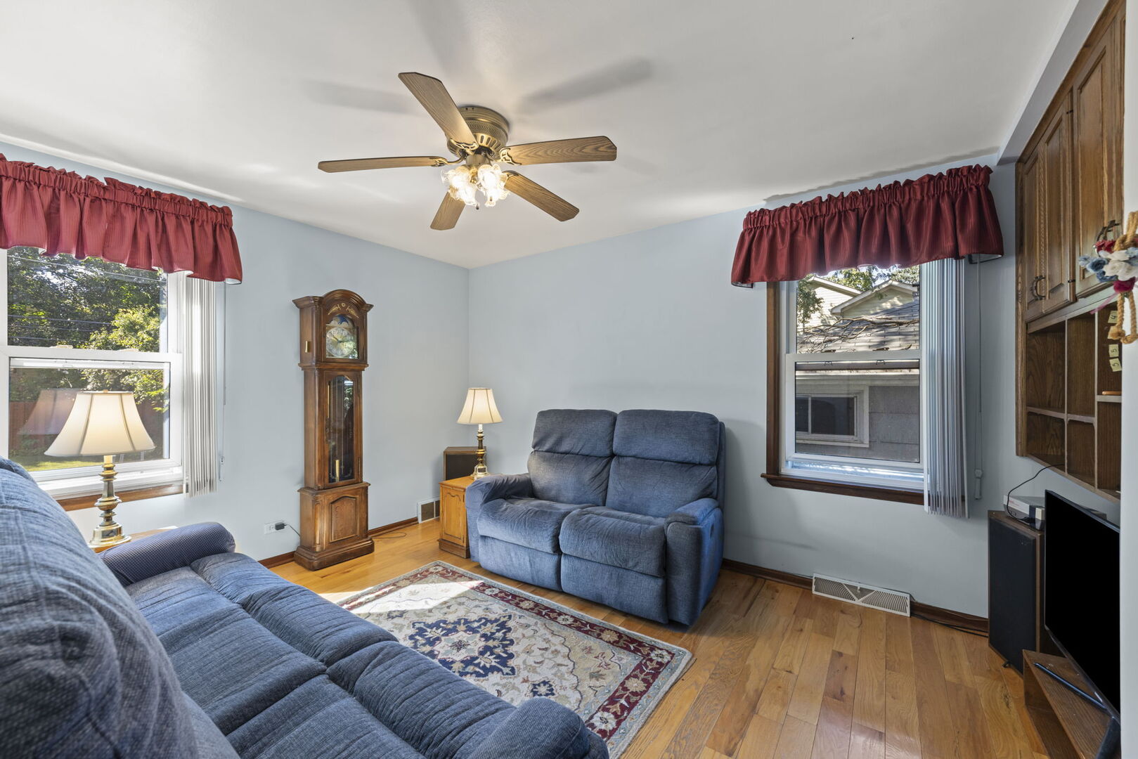 Living room with blue sofas, hardwood floors, a grandfather clock, and burgundy window valances.