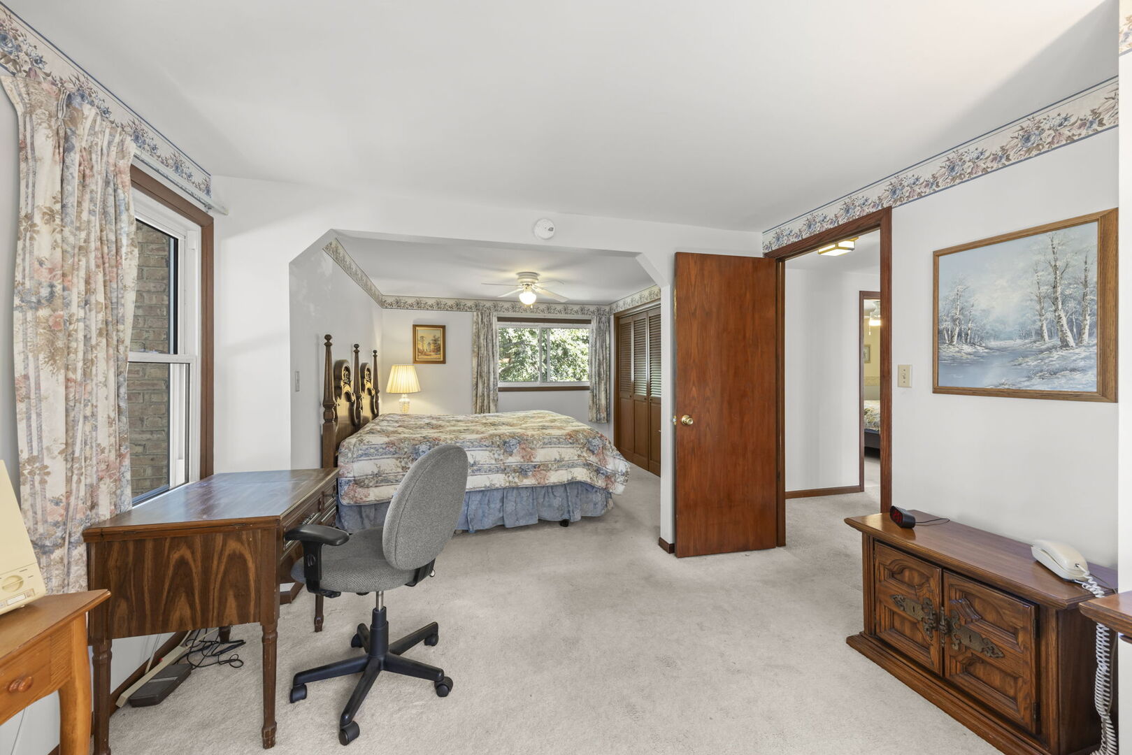 Bedroom with a floral-patterned bed, large window, desk, and dark wood furniture.