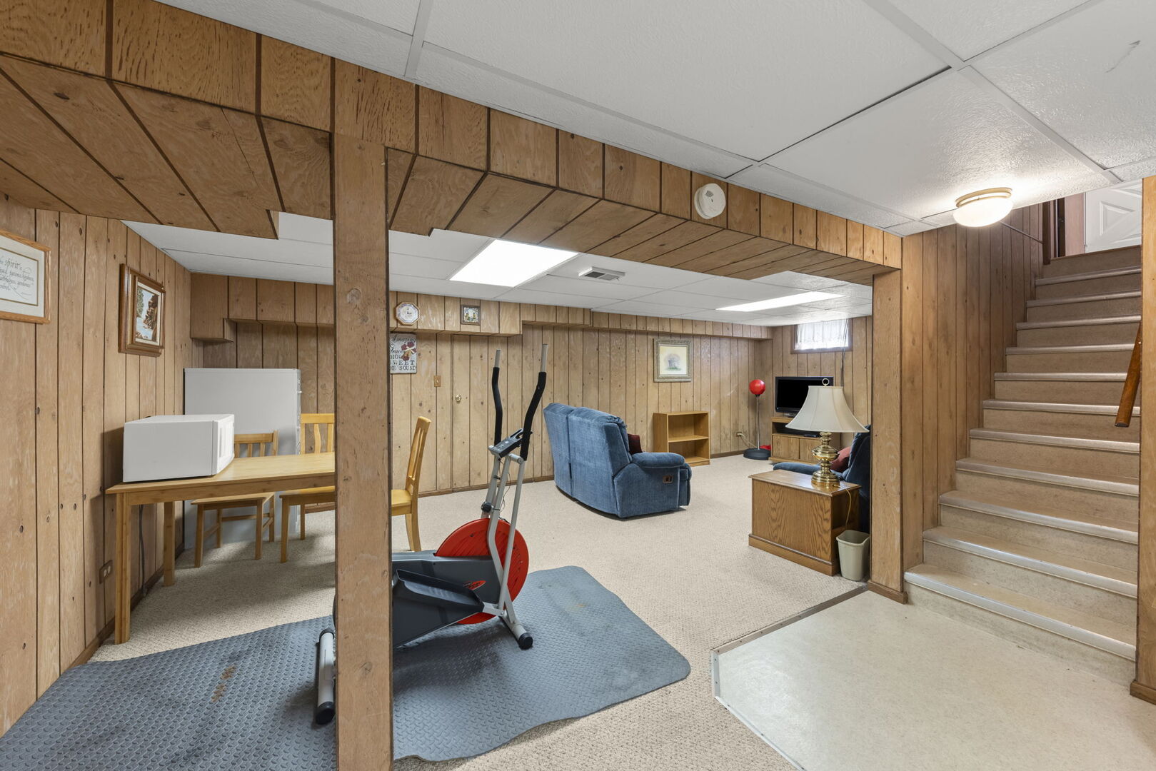 Basement recreation room with wood paneling, exercise equipment, blue seating, and carpeted stairs.