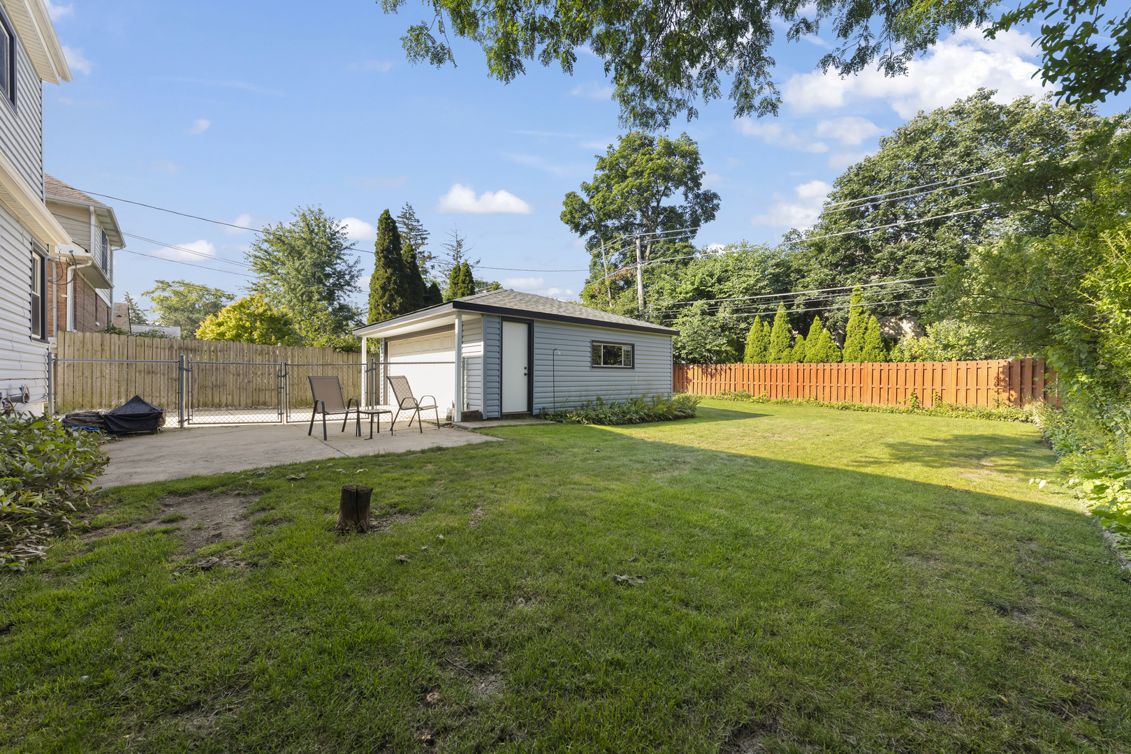 Backyard lawn and fenced patio with outdoor chairs beside a detached garage.