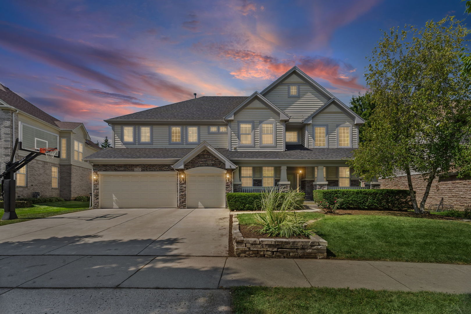 Two-story gray home with stone accents, a three-car garage, and a basketball hoop at dusk.