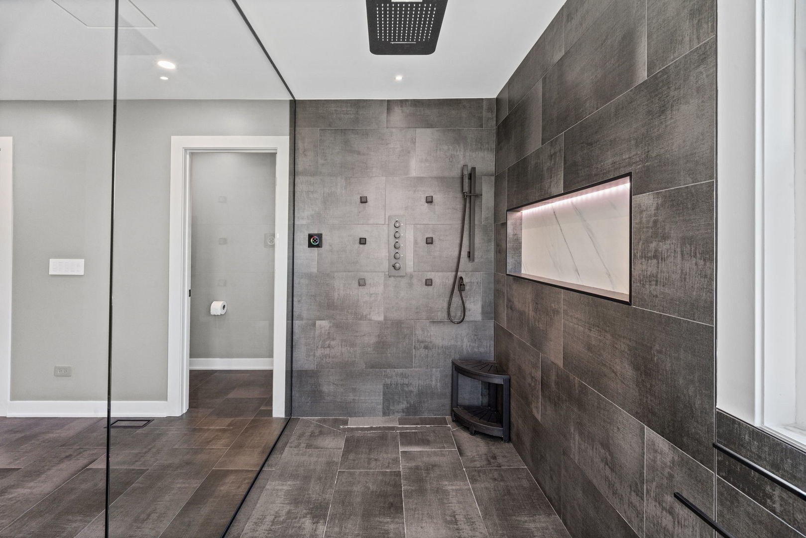 Walk-in shower with dark gray tile, multiple shower controls, a lit wall niche, and a corner seat.