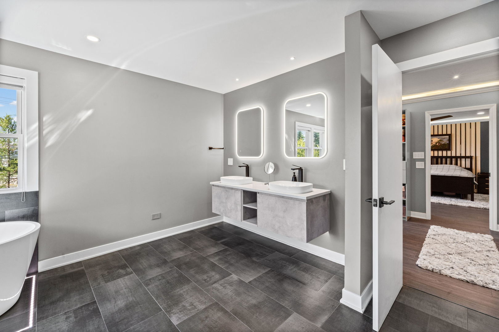 Modern bathroom with a floating double vanity, illuminated mirrors, dark tile, and a freestanding tub.