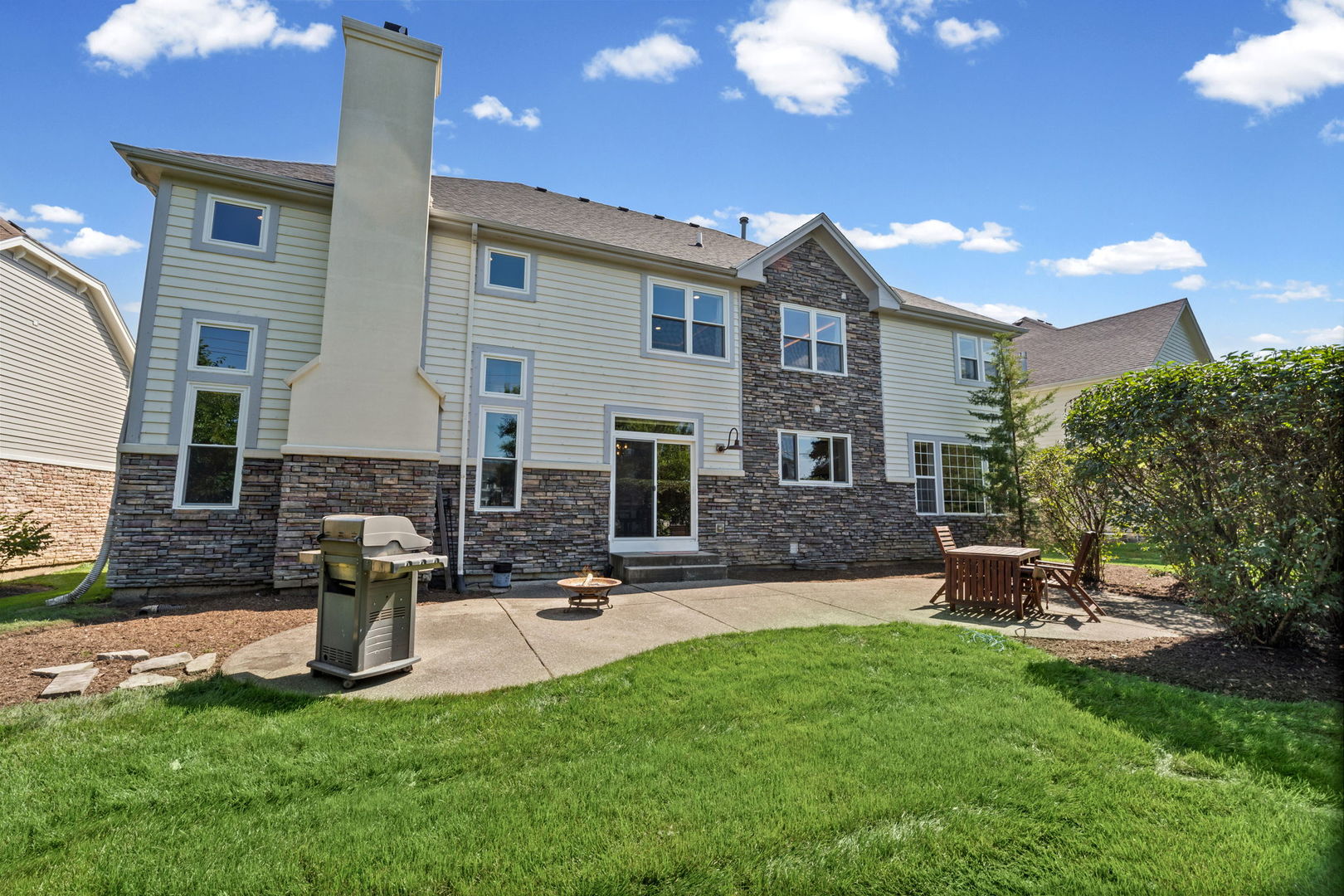 Rear of a two-story home with stone veneer, a tall chimney, and a patio with grill and seating.