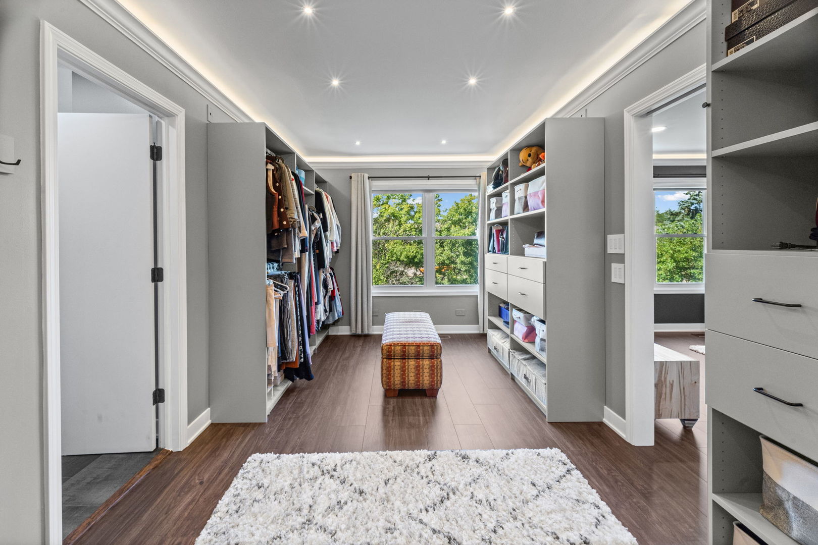 Walk-in closet with gray built-ins, hanging clothes, a central bench, and a large window overlooking trees.