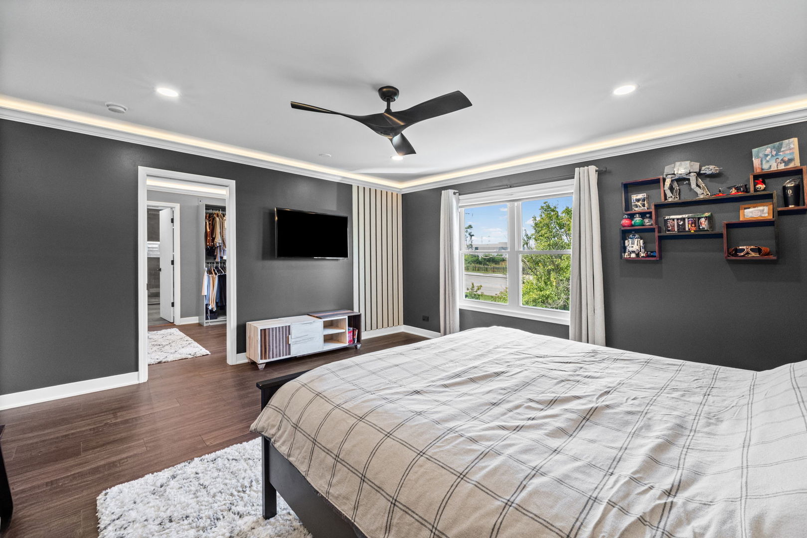 Bedroom with dark gray walls, a plaid-covered bed, display shelves, and a window overlooking greenery.