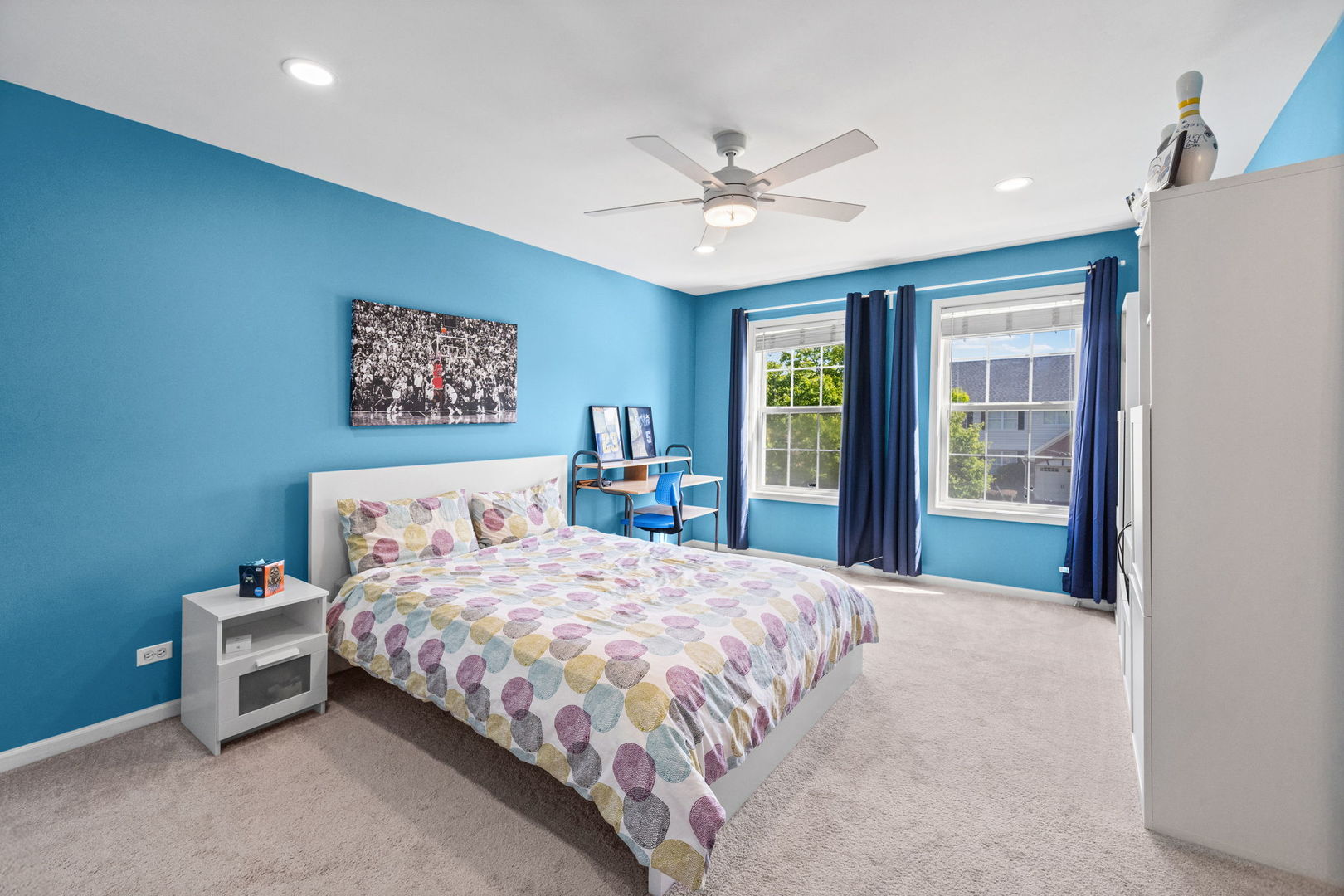 Furnished bedroom with bright blue walls, patterned bedding, and two curtained windows.