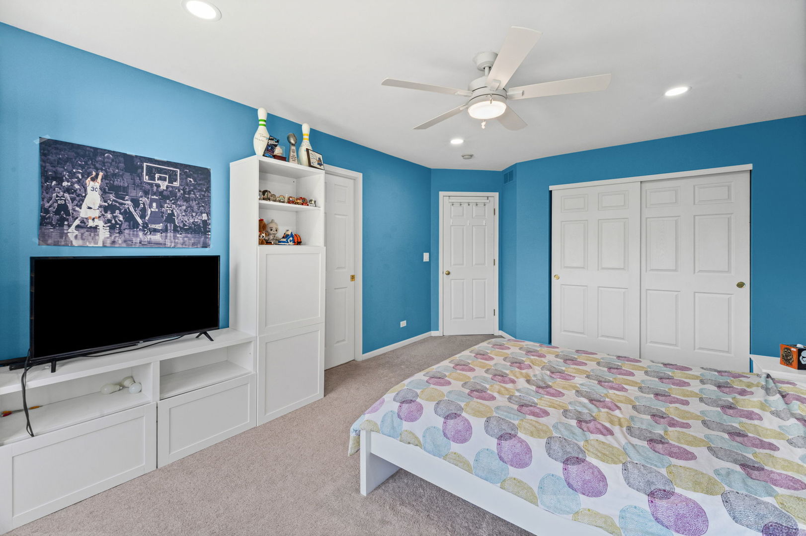 Blue-walled bedroom with built-in white storage, sliding closet doors, ceiling fan, and patterned bedspread.
