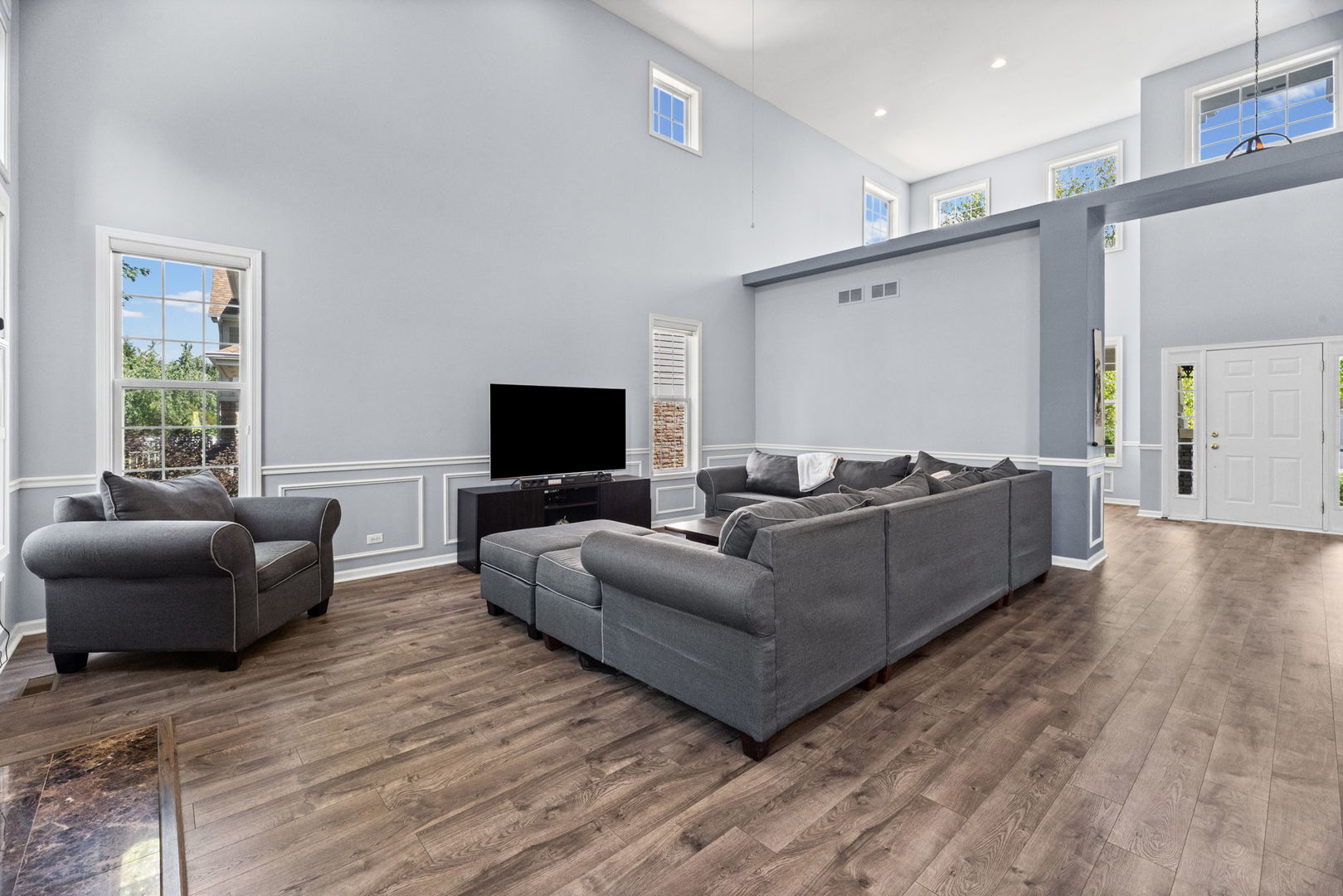 Furnished living room with vaulted ceiling, gray seating, large windows, and wood-look floors.