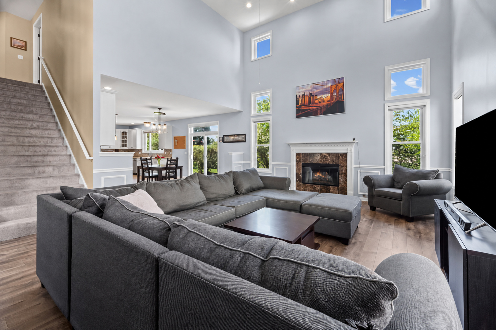 Double-height living room with a gray sectional, fireplace, and staircase beside an open kitchen.