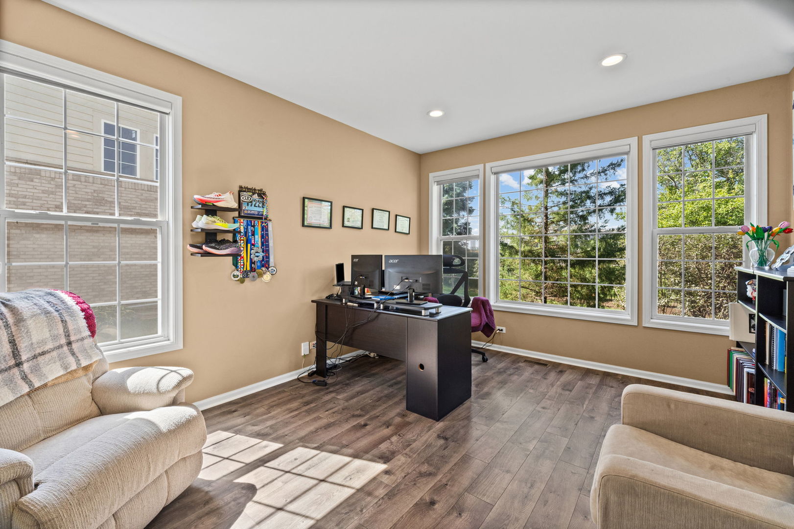 Home office with a dark desk, dual monitors, large windows, and upholstered seating.
