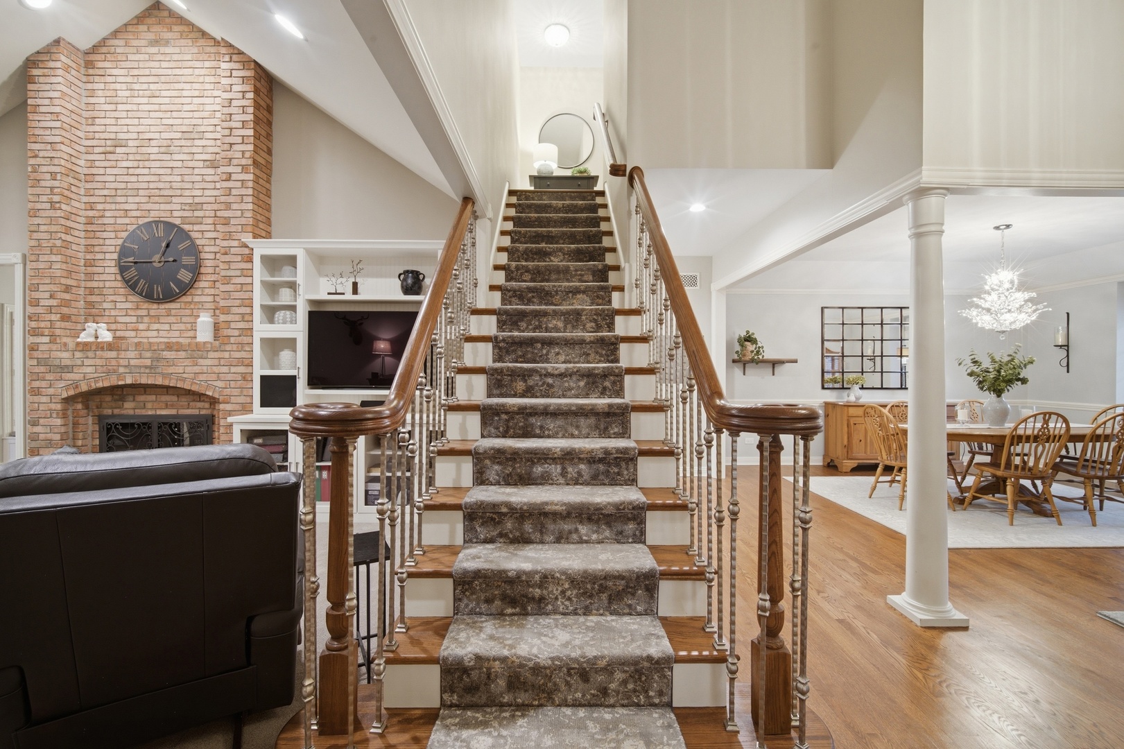 Carpeted staircase with wood handrails beside a tall brick fireplace and open dining area.