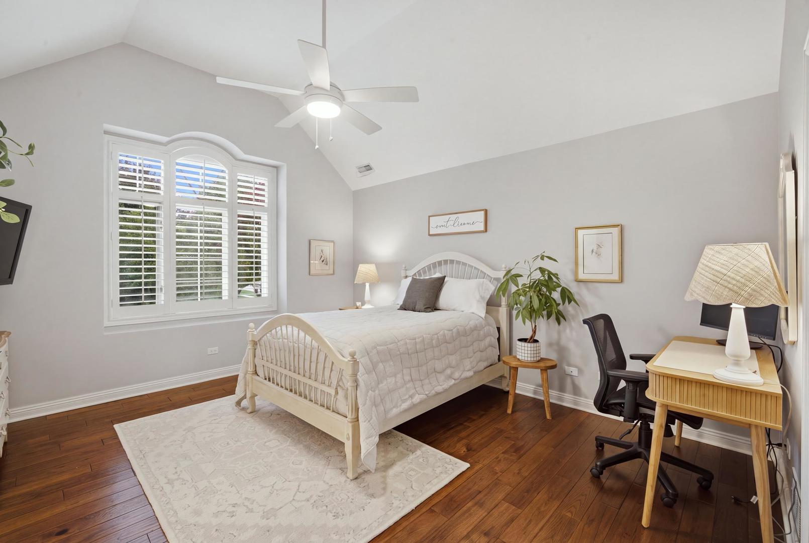 Furnished bedroom with a white bed, vaulted ceiling, plantation-shuttered window, and desk.