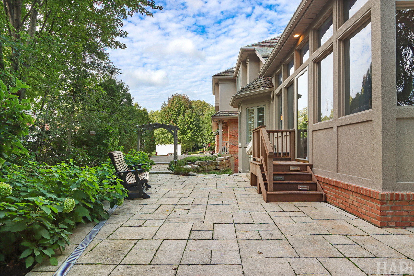 Stone-paved patio beside a home, with wooden steps, a dark chair, and lush garden beds.