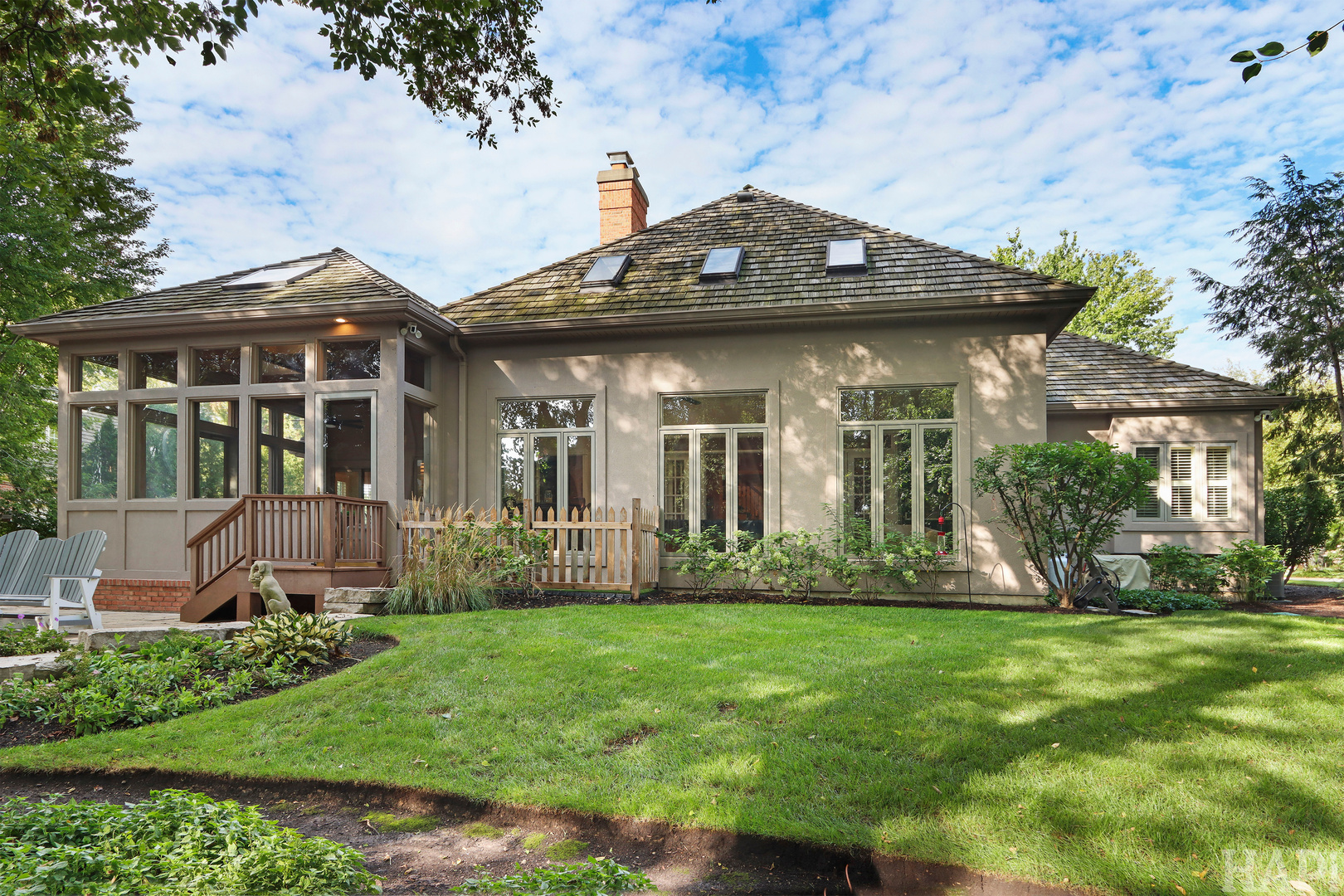 Rear of a beige house with a glass sunroom, skylights, deck steps, and a green lawn.
