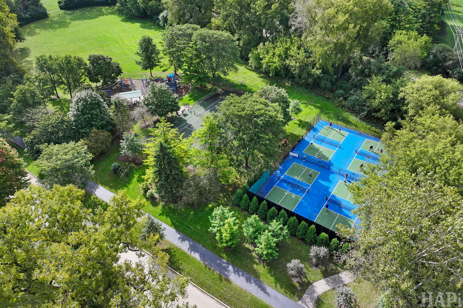 Aerial view of a tree-lined park with blue pickleball courts, a playground, and walking paths.
