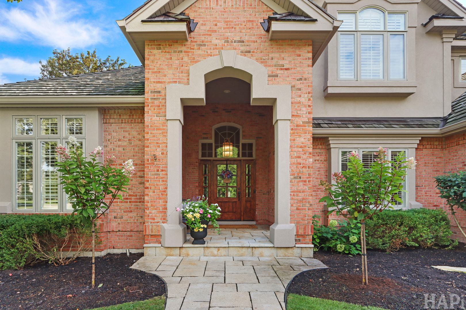 Brick home with a covered entry, wood front door, stone walkway, and flowering landscaping.
