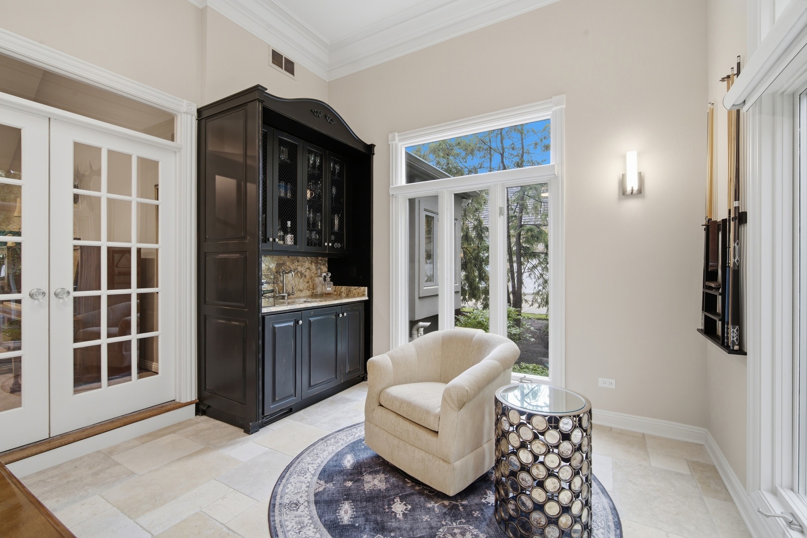 Sitting area with a dark built-in wet bar, cream armchair, and large windows overlooking trees.