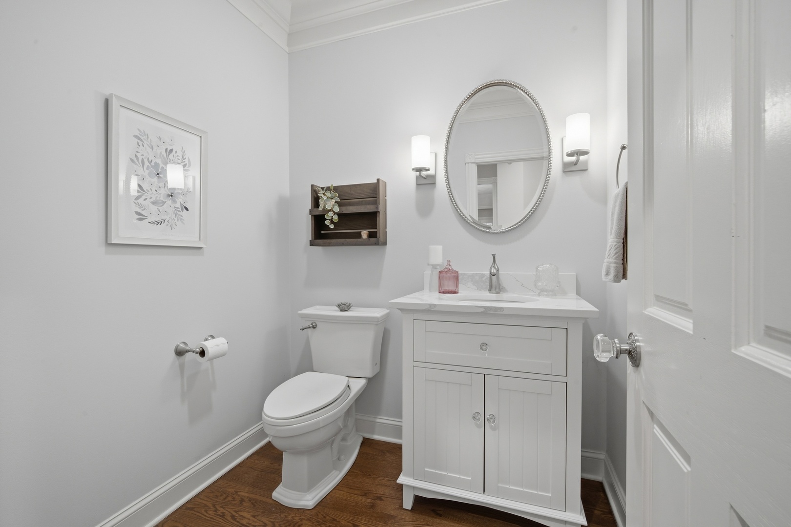 Bathroom with a white vanity, oval mirror, wall sconces, and dark wood flooring.