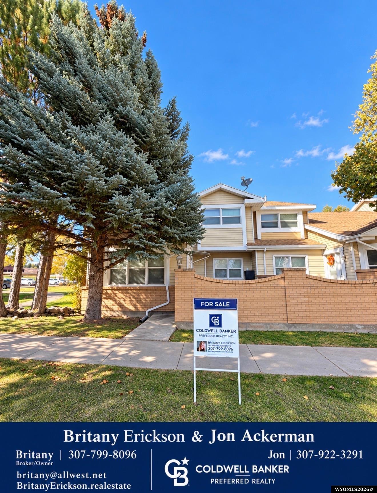 Beige residence behind a brick wall, with a large evergreen and Coldwell Banker for-sale sign.