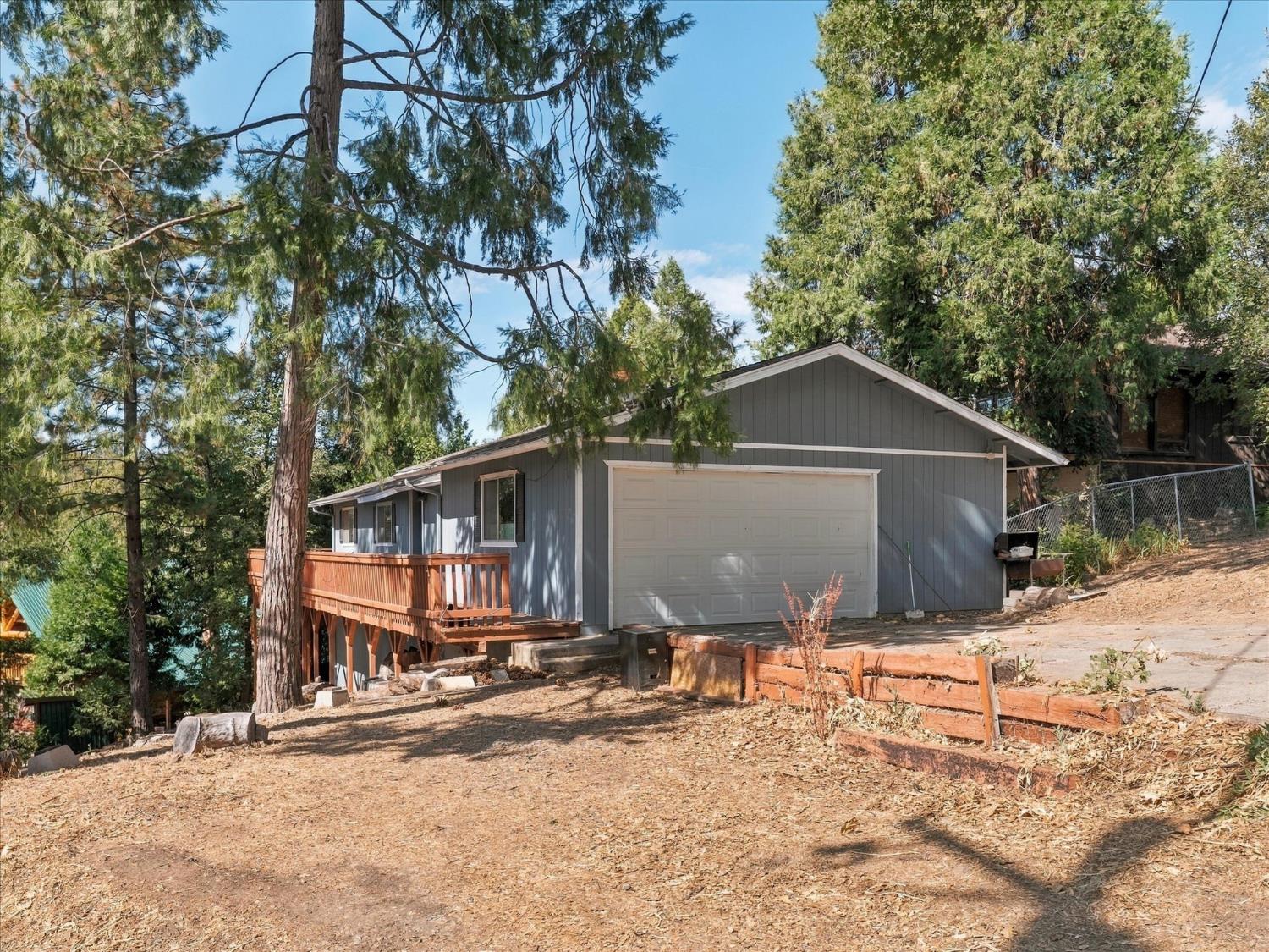 Gray single-story house with a white double garage door and raised wooden deck among tall trees.