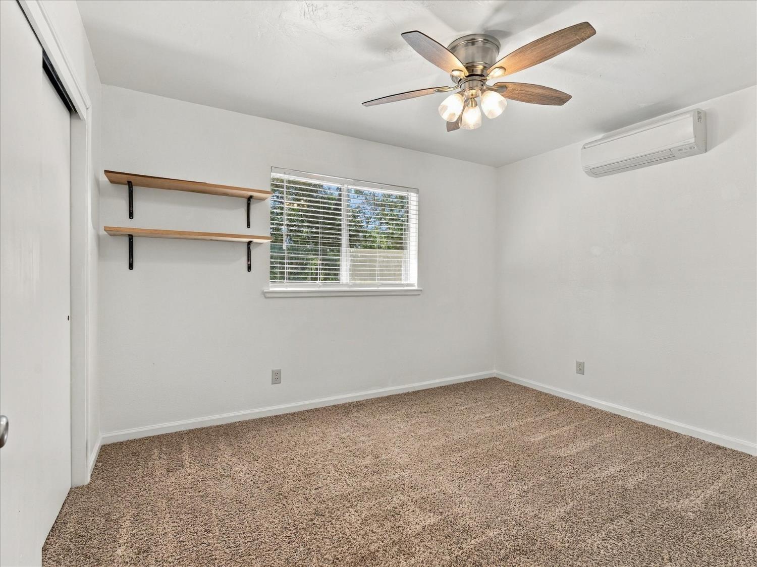 Empty carpeted room with wooden shelves, a window, ceiling fan, and wall-mounted air conditioner.