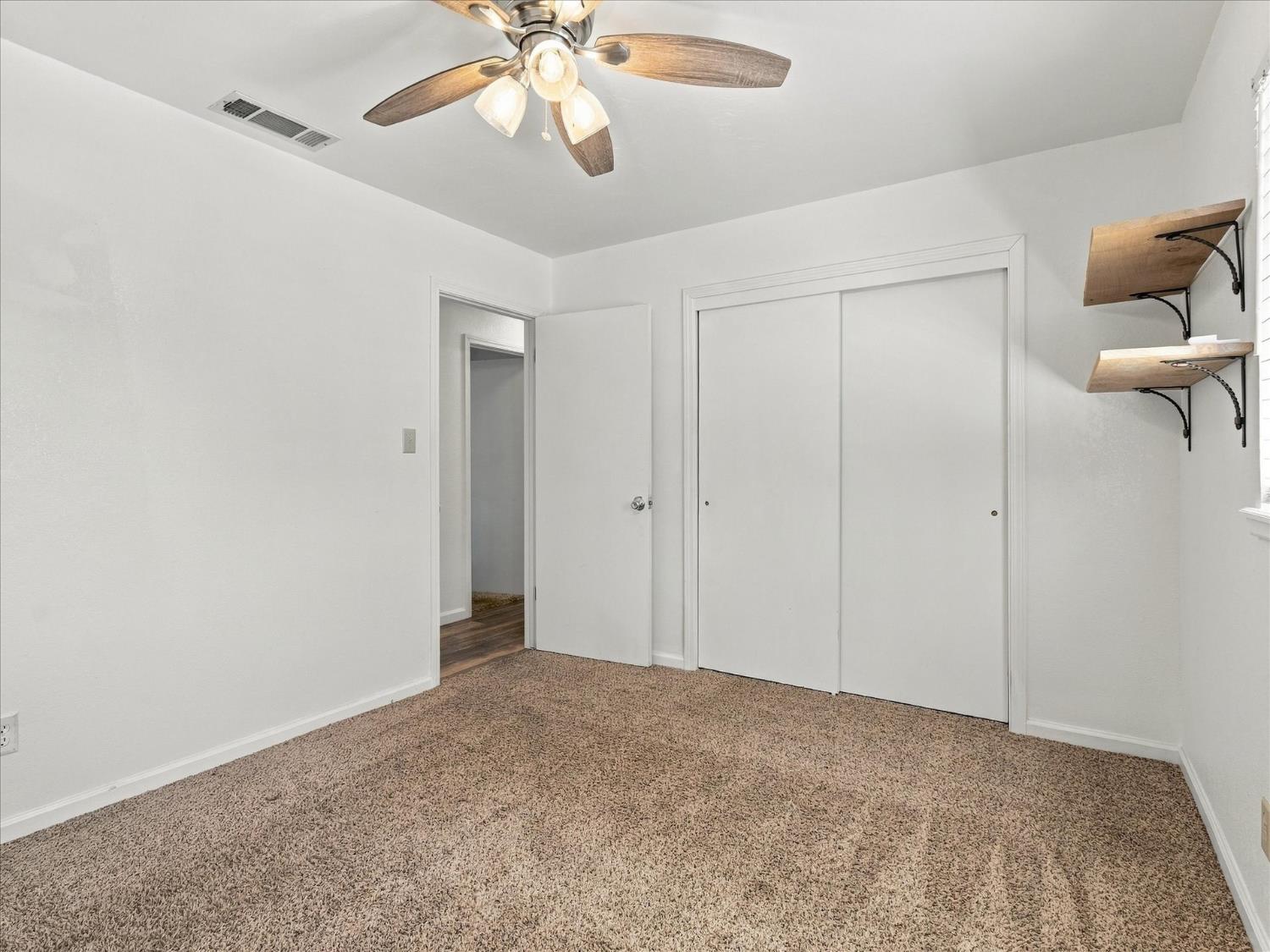 Empty carpeted bedroom with sliding closet doors, a ceiling fan, and wooden wall shelves.