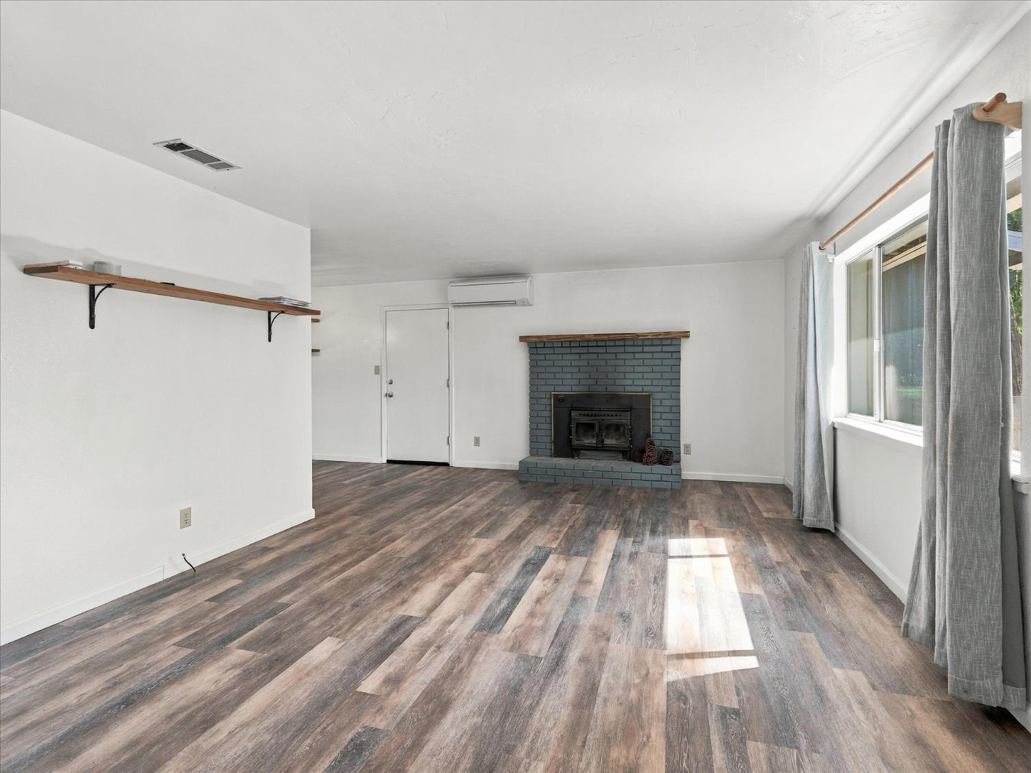 Empty living room with a blue brick fireplace, wood-burning stove, and large curtained windows.