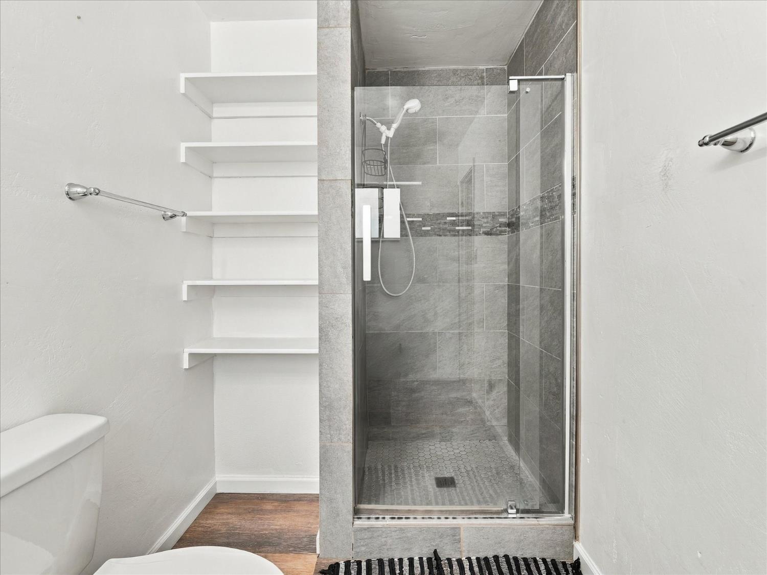 Bathroom with a gray tiled glass shower and open white wall shelves.