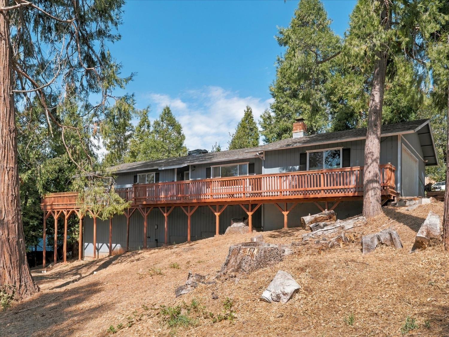 Gray house with a long raised wooden deck on a dry, sloping lot among tall trees.