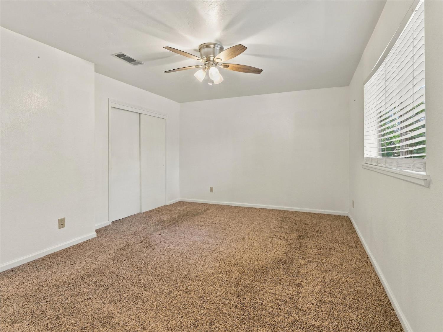 Empty carpeted bedroom with a sliding-door closet, window blinds, and ceiling fan.