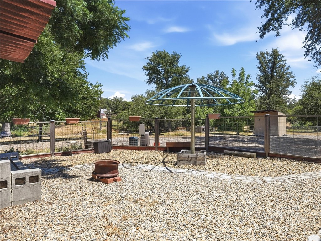 Fenced gravel yard with a circular canopy frame, fire pit, raised planters, and mature trees.