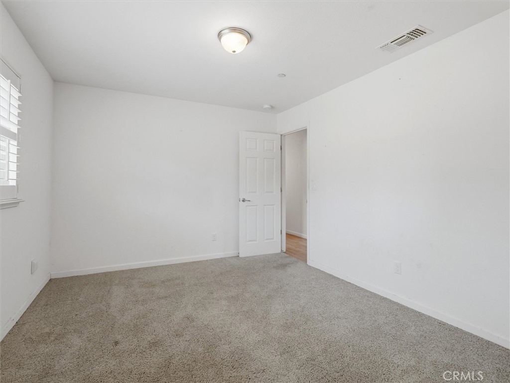 Empty bedroom with gray carpet, window blinds, and an open door to a wood-floored hallway.