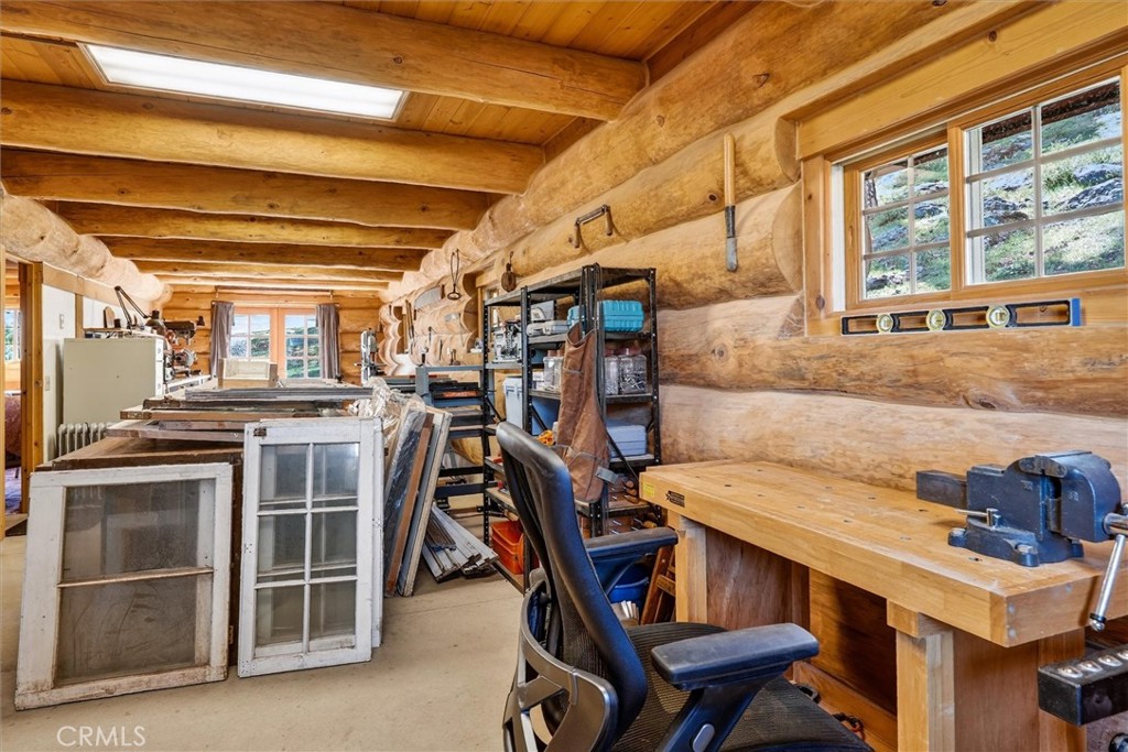 Log-walled workshop with workbenches, tools, and windows overlooking a rocky hillside.