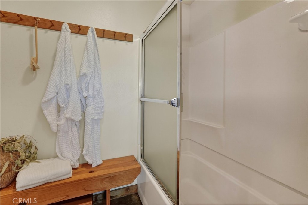 Bathroom shower with glass door, wooden bench, hanging white robes, and folded towels.