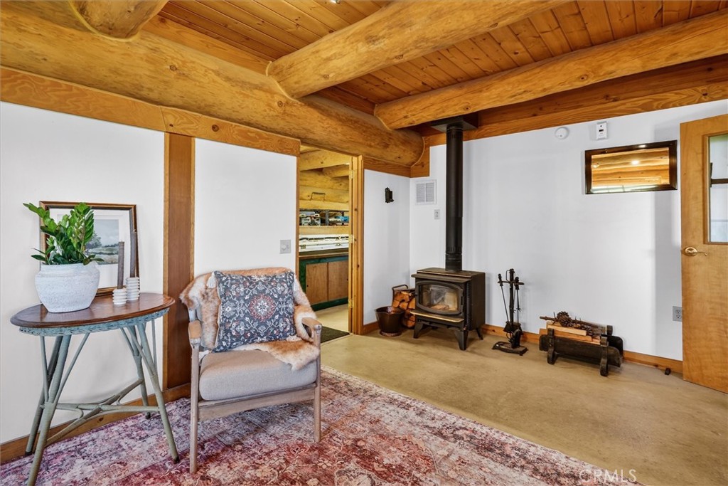 Living room with exposed log beams, a wood-burning stove, patterned rug, and doorway to the kitchen.